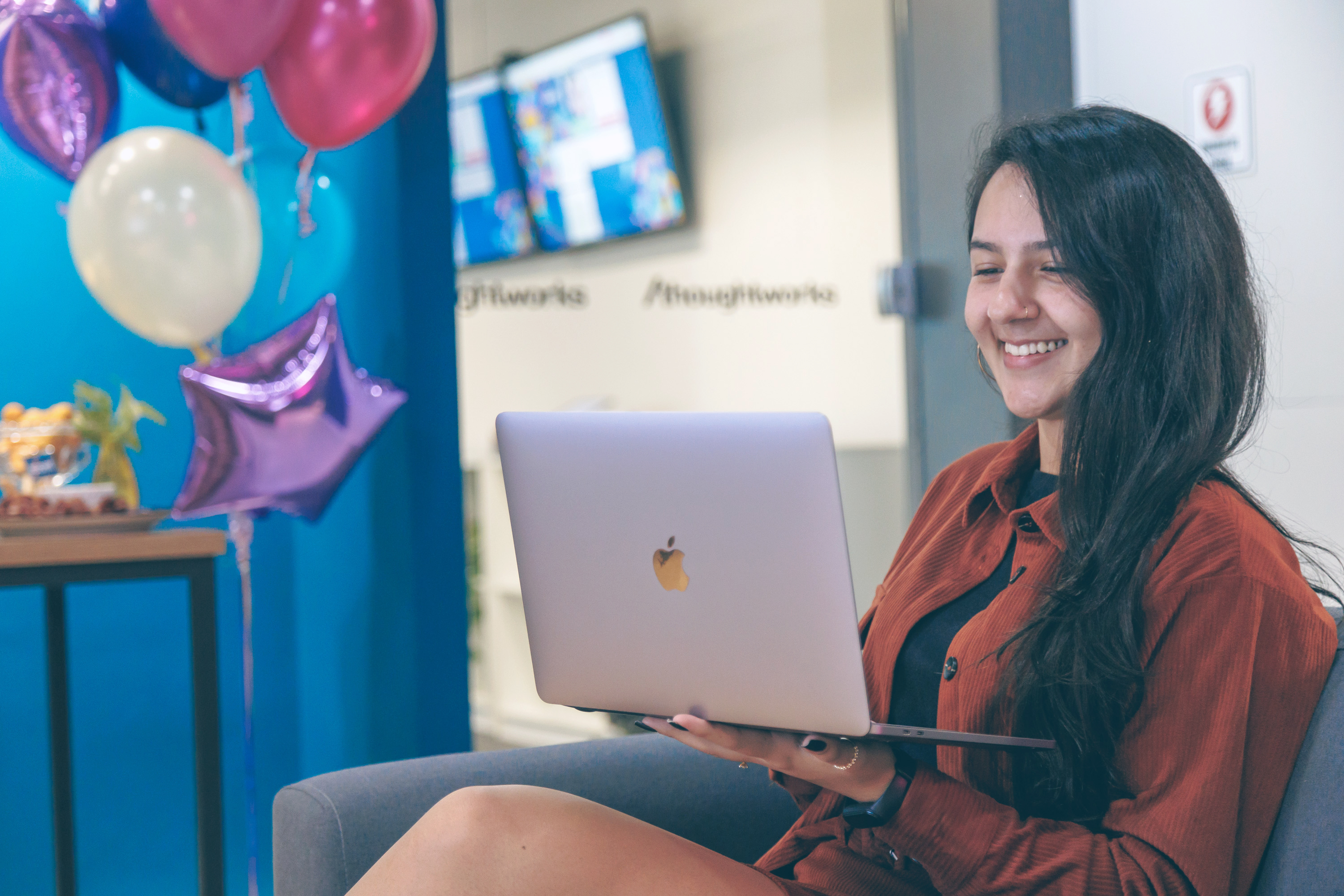 Woman smiling and sitting on a gray sofa with a laptop on her lap. In the background of the image, there is the Thoughtworks office in Porto Alegre, with balloons and a table on her left side.
