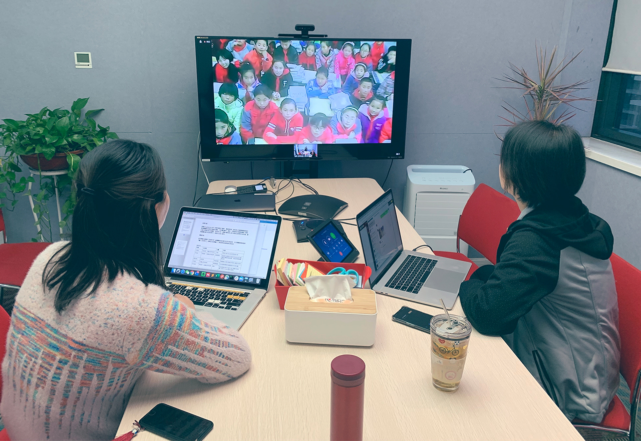 Duas Thoughtworkers em uma mesa, conectadas por vídeo com um grande grupo de crianças, na China rural, sentadas no chão ouvindo.