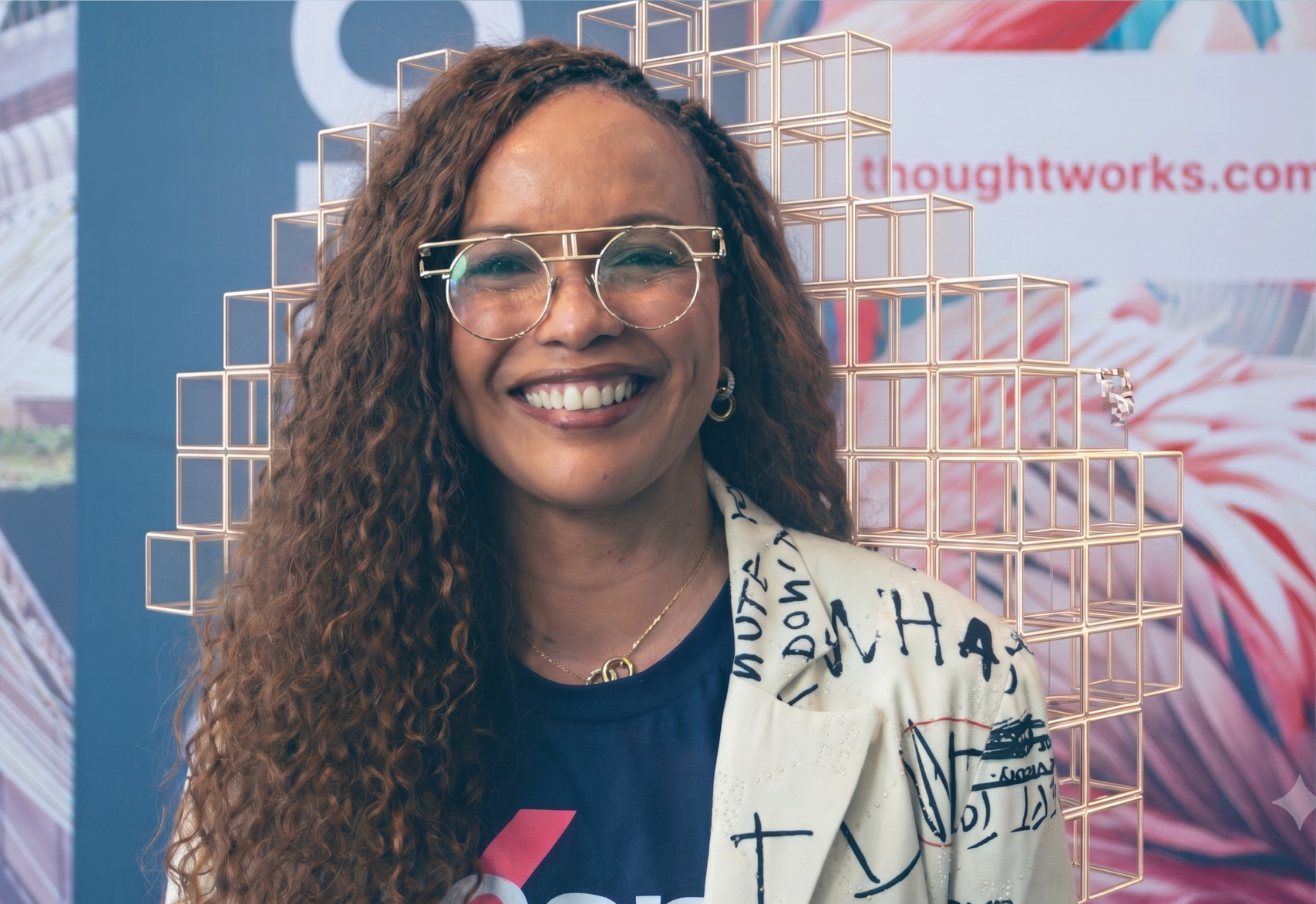 A photograph of one of our DEI leaders, wearing a woolen hat which she is holding on her head. She is smiling and her blue hair matches the mural on the wall beehind her, which is mingled with AI cubes.