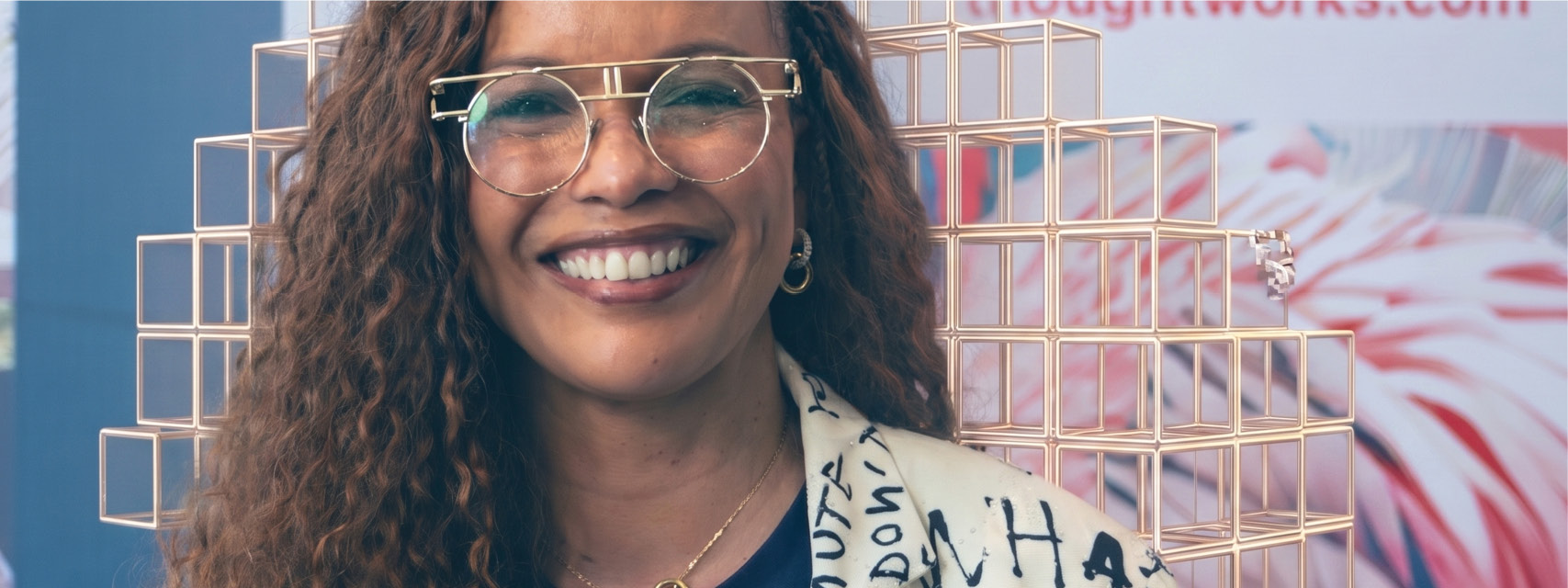 A photograph of one of our DEI leaders, wearing a woolen hat which she is holding on her head. She is smiling and her blue hair matches the mural on the wall beehind her, which is mingled with AI cubes.