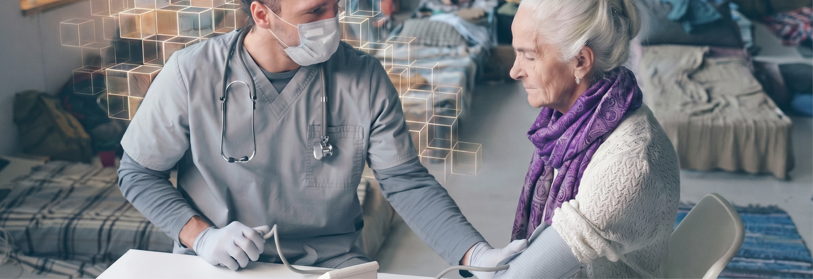 A young male medic is checking an older ladys blood pressure. They are sitting at a table in the foreground, both in grey but she has a purple scarf on. In the background are many small beds, with a group of people sitting / resting on them.