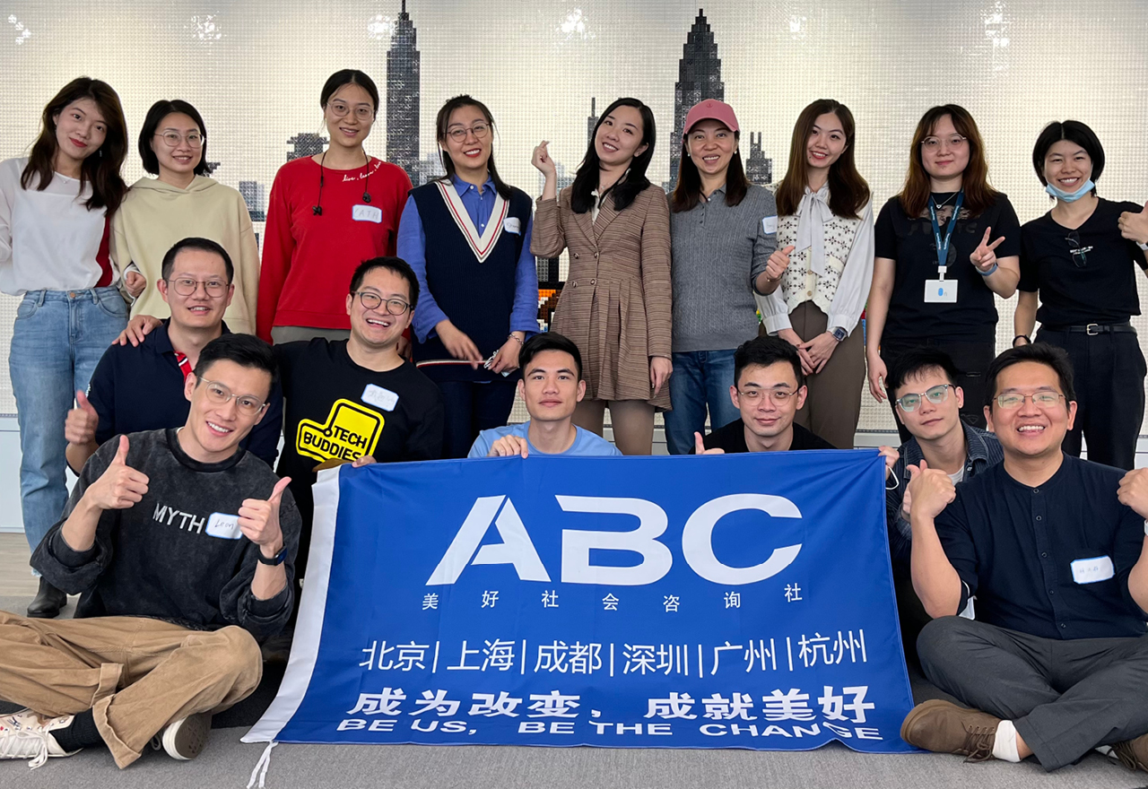 A photograph of a group of individuals gathered around the ABC logo flag, some are smiling and posing with hand gestures for the photographer 