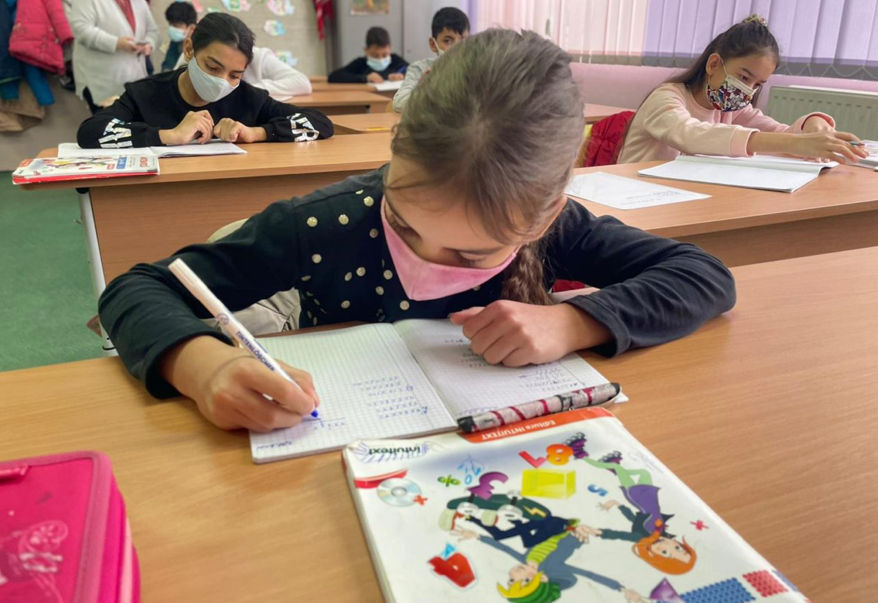 A photograph of a young girl sitting at her desk writing at school, with her classmates in the background.