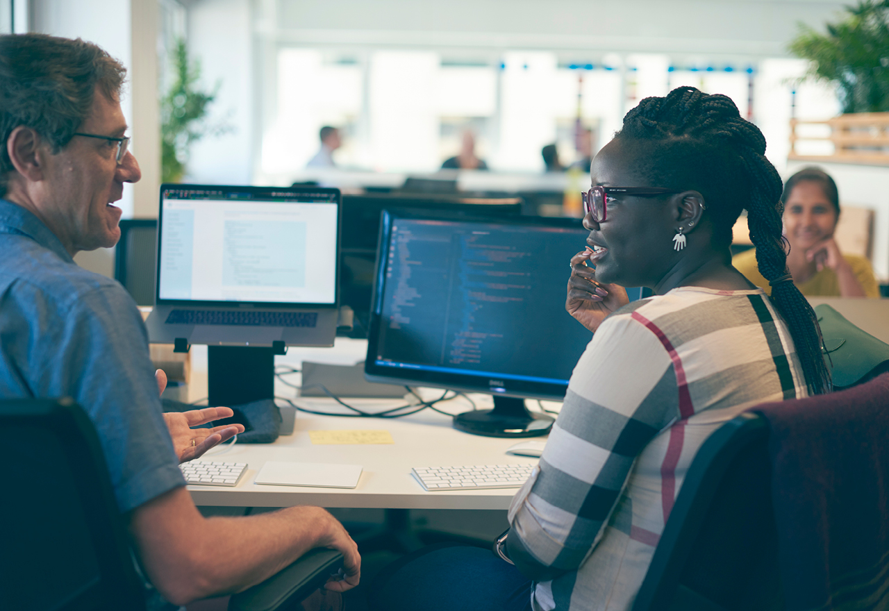 two Thoughtworkers sat in front of a desk with two computers having a conversation 