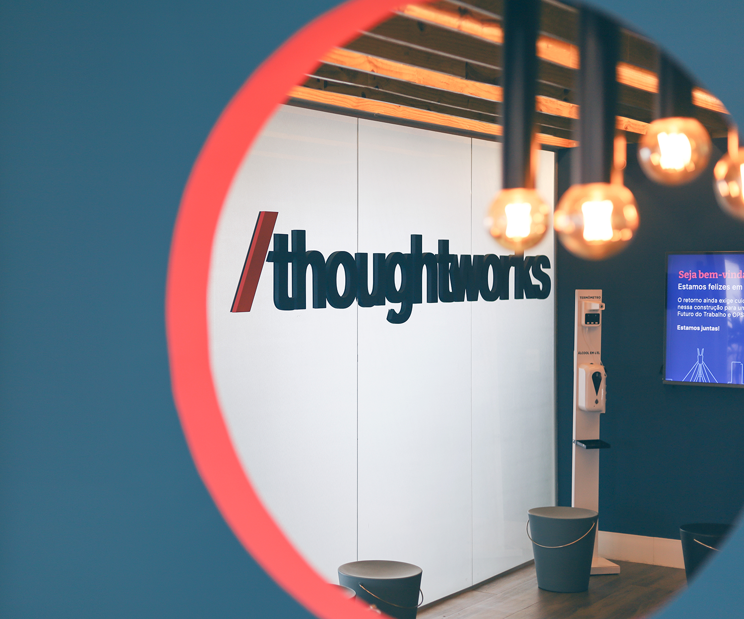 Imagem do escritório da Thoughtworks em Recife, mostrando a recepção com o logo na parede, uma televisão e luzes iluminando o ambiente.