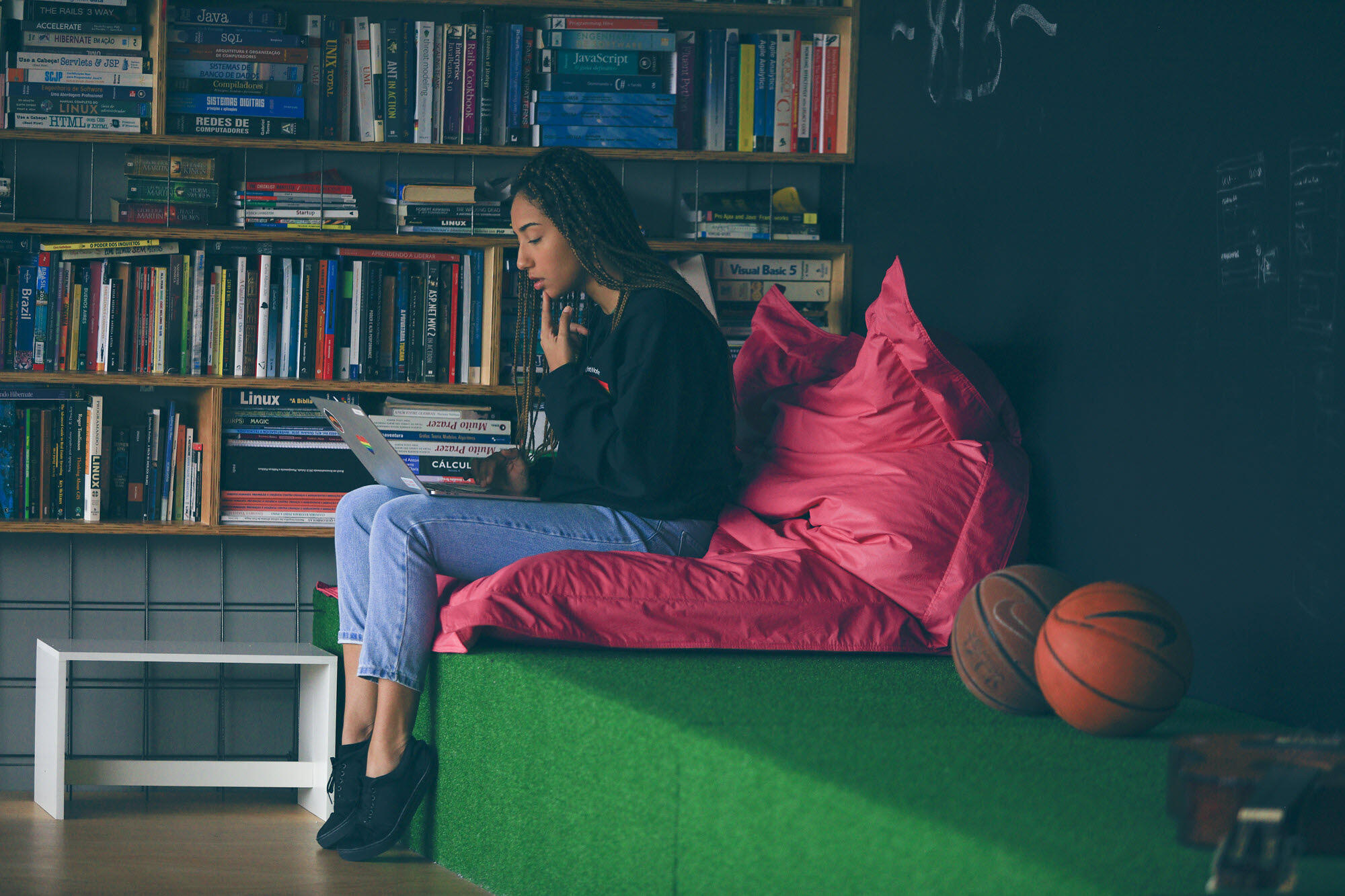 Uma jovem mulher na frente de uma grande estante com vários livros, sentada em um banco e uma grande almofada trabalhando em um laptop.