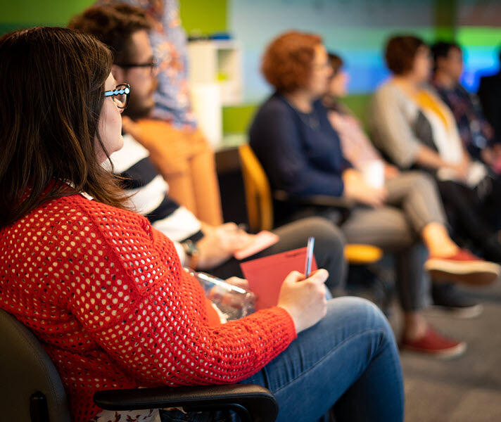 Imagem do perfil de um grupo de Thoughtworkers assistindo uma apresentação. Uma mulher está mais próxima da câmera em foco.