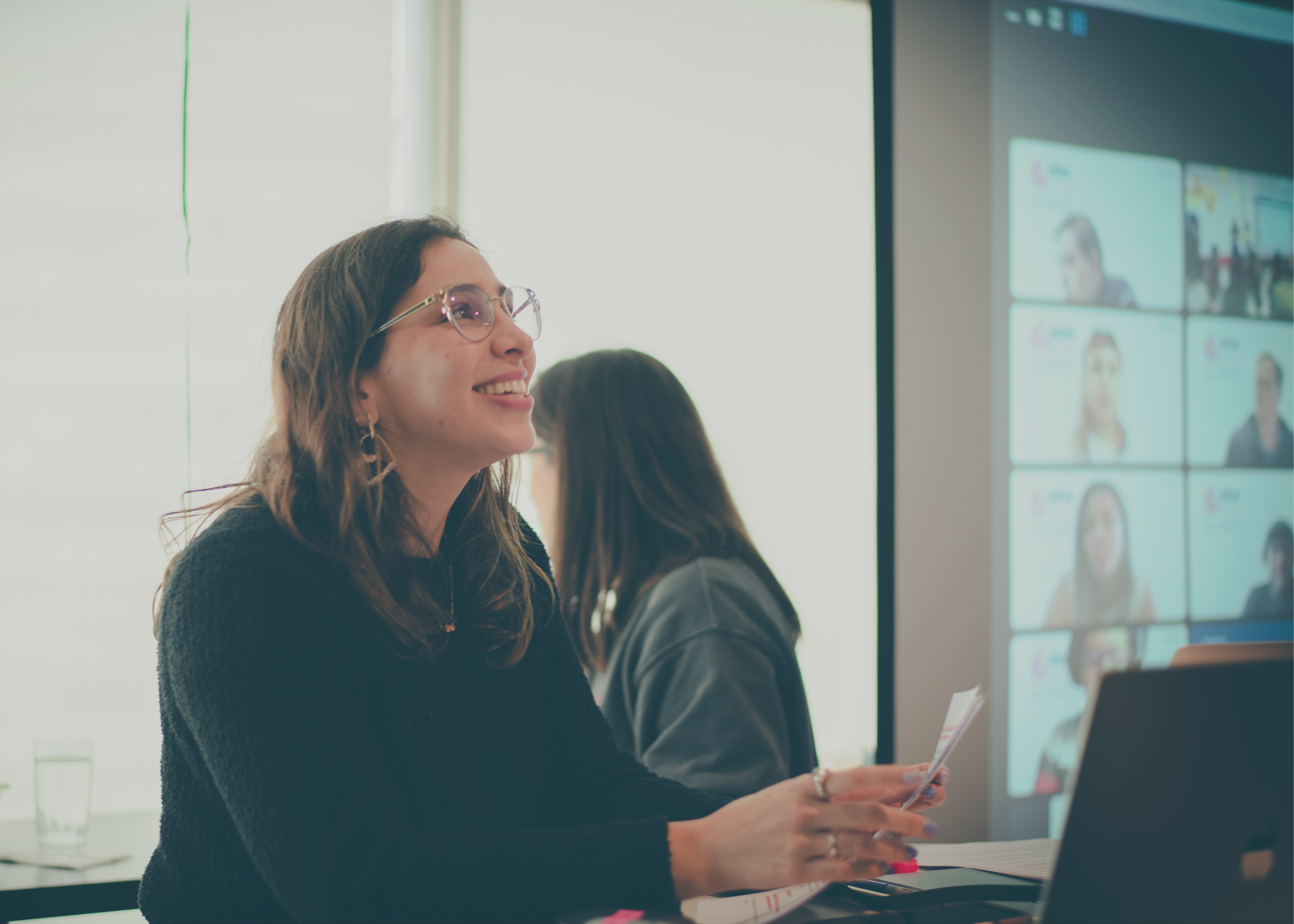 Una mujer, sonriendo mientras dirige su mirada hacia arriba, toma parte en una reunión dentro de un salón de eventos. En segundo plano, una pantalla proyecta una reunión de Zoom con la presencia de otras personas.