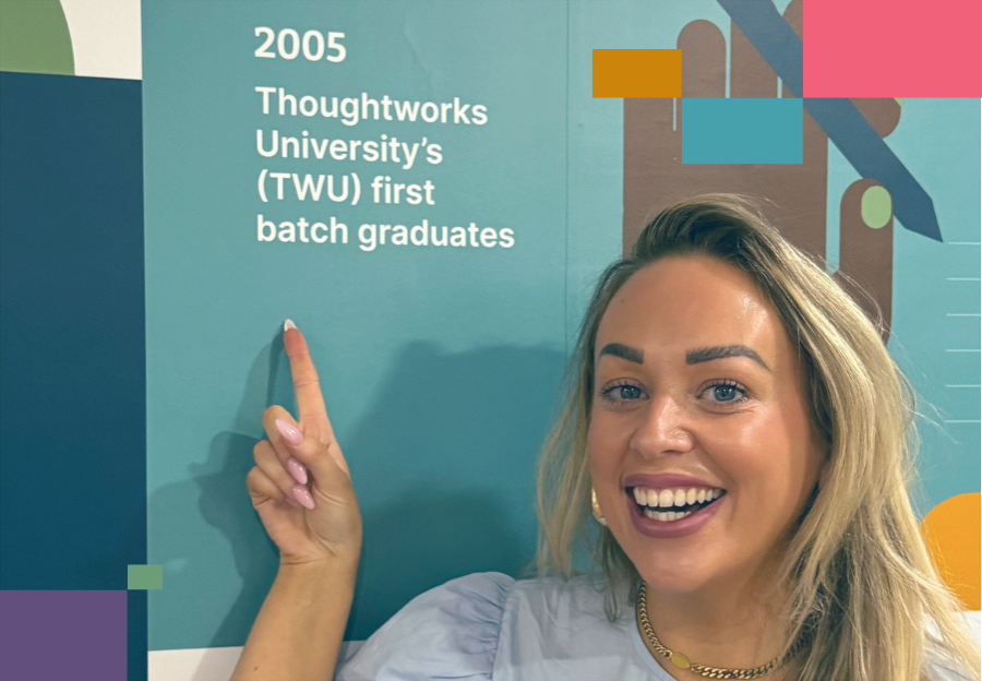 Jade, Global Head of First Year Experience at Thoughtworks, smiling and pointing to a wall graphic highlighting the 2005 milestone: “Thoughtworks University’s (TWU) first batch graduates,” during her visit to the India office. Jade, Global Head of First Year Experience at Thoughtworks, smiling and pointing to a wall graphic highlighting the 2005 milestone: “Thoughtworks University’s (TWU) first batch graduates,” during her visit to the India office.