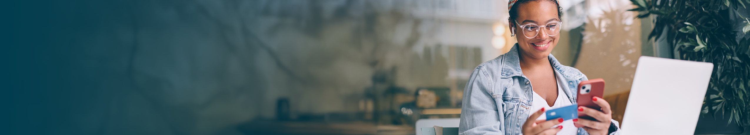 Smiling young woman with curly hair and glasses holding a red smartphone and a blue credit card, seated in a casual setting with a laptop in front of her, engaged in an online transaction.