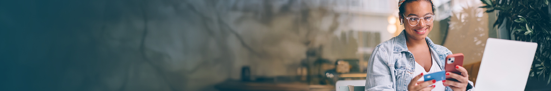 Smiling young woman with curly hair and glasses holding a red smartphone and a blue credit card, seated in a casual setting with a laptop in front of her, engaged in an online transaction.