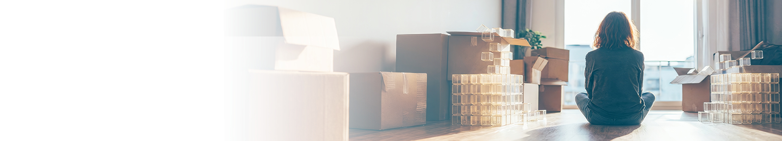 A person sits cross-legged on a wooden floor, surrounded by cardboard moving boxes and transparent plastic containers, gazing out a large sunlit window. The left side of the image fades to white, offering space for text or design elements.