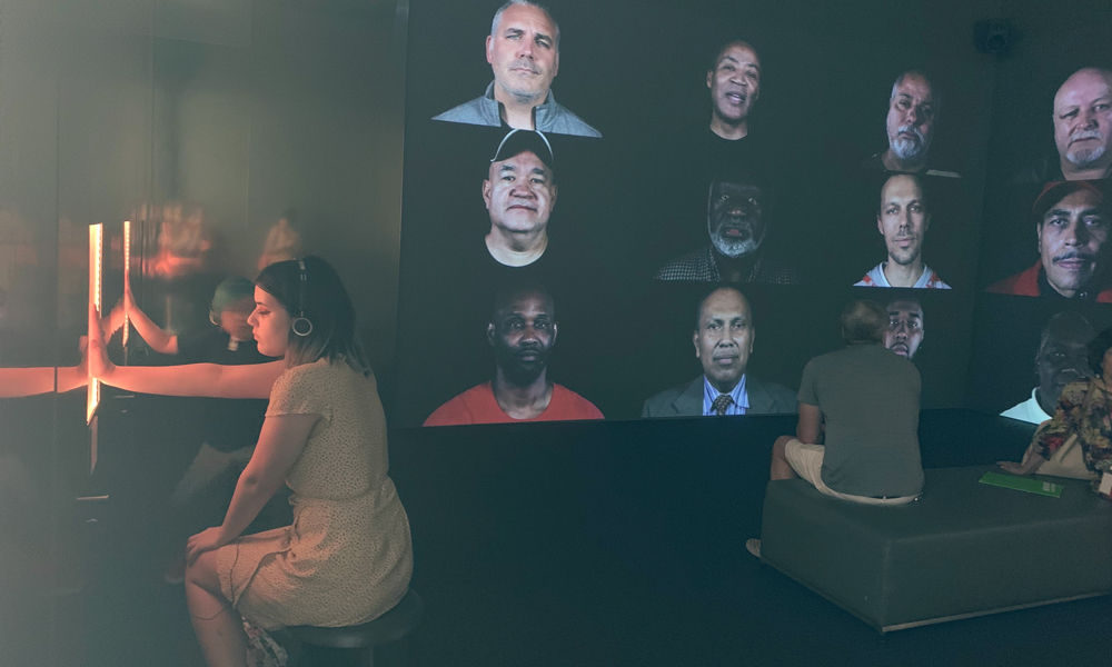 Chica con la mano en una instalación de arte, y una pared de fotografías en el fondo