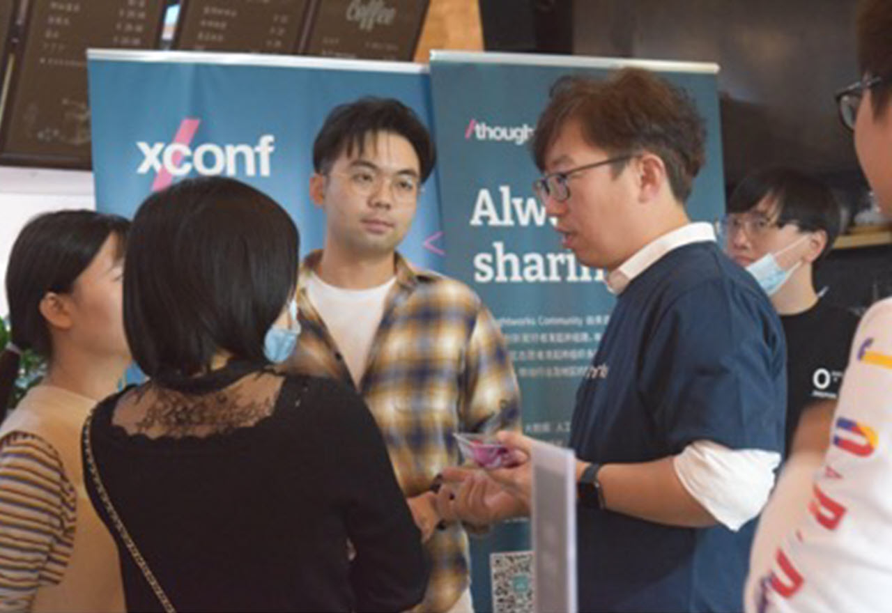 Group of two women and two men stand in a circle talking. Two other people look on. XConf and Thoughtworks branded pop-up banners in the background.