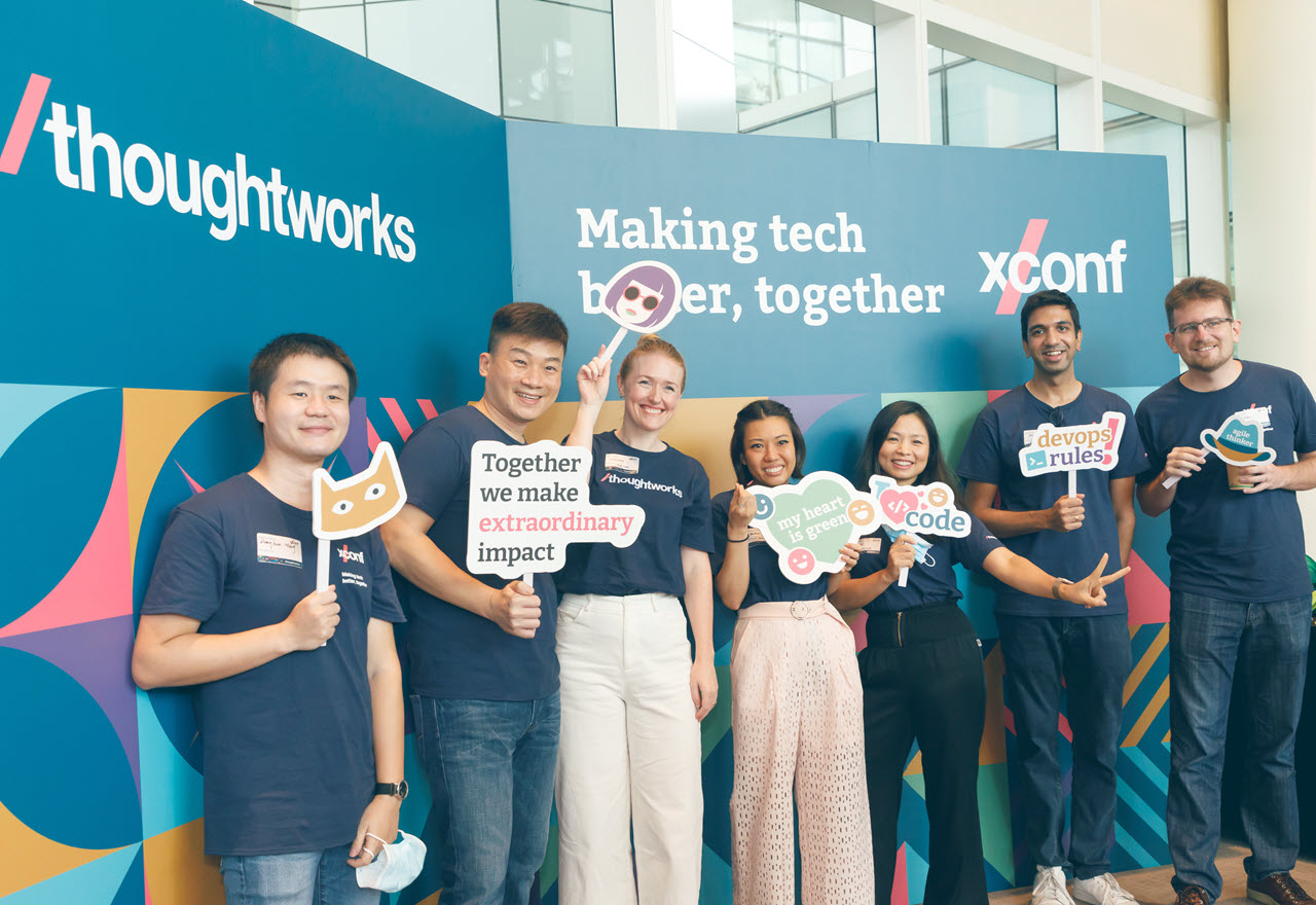 Group of XConf Singapore 2022 event speakers and organizers standing in front of a branded backdrop with each person holding up a fun photo prop sign, assorted sayings and images