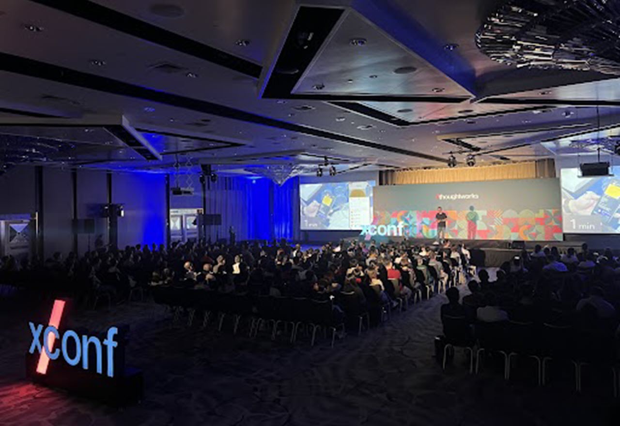 A view of the XConf Singapore branded stage from behind the rows of the seated audience and a lit up XConf logo sign.