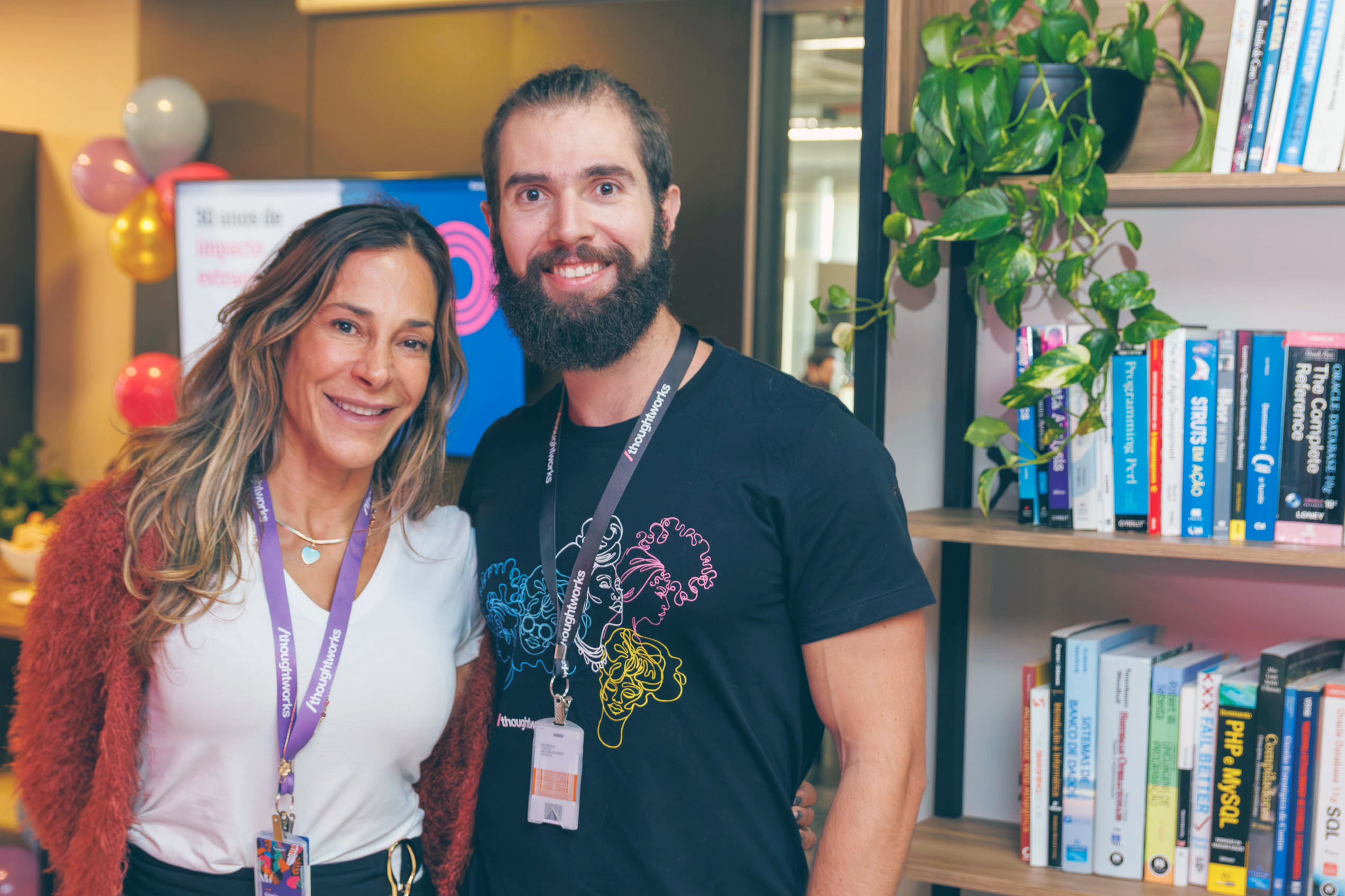 A woman on the left and a man embracing with badges in the Thoughtworks office. To the left of the man, there is a bookshelf. In the background, there is a television and balloons.