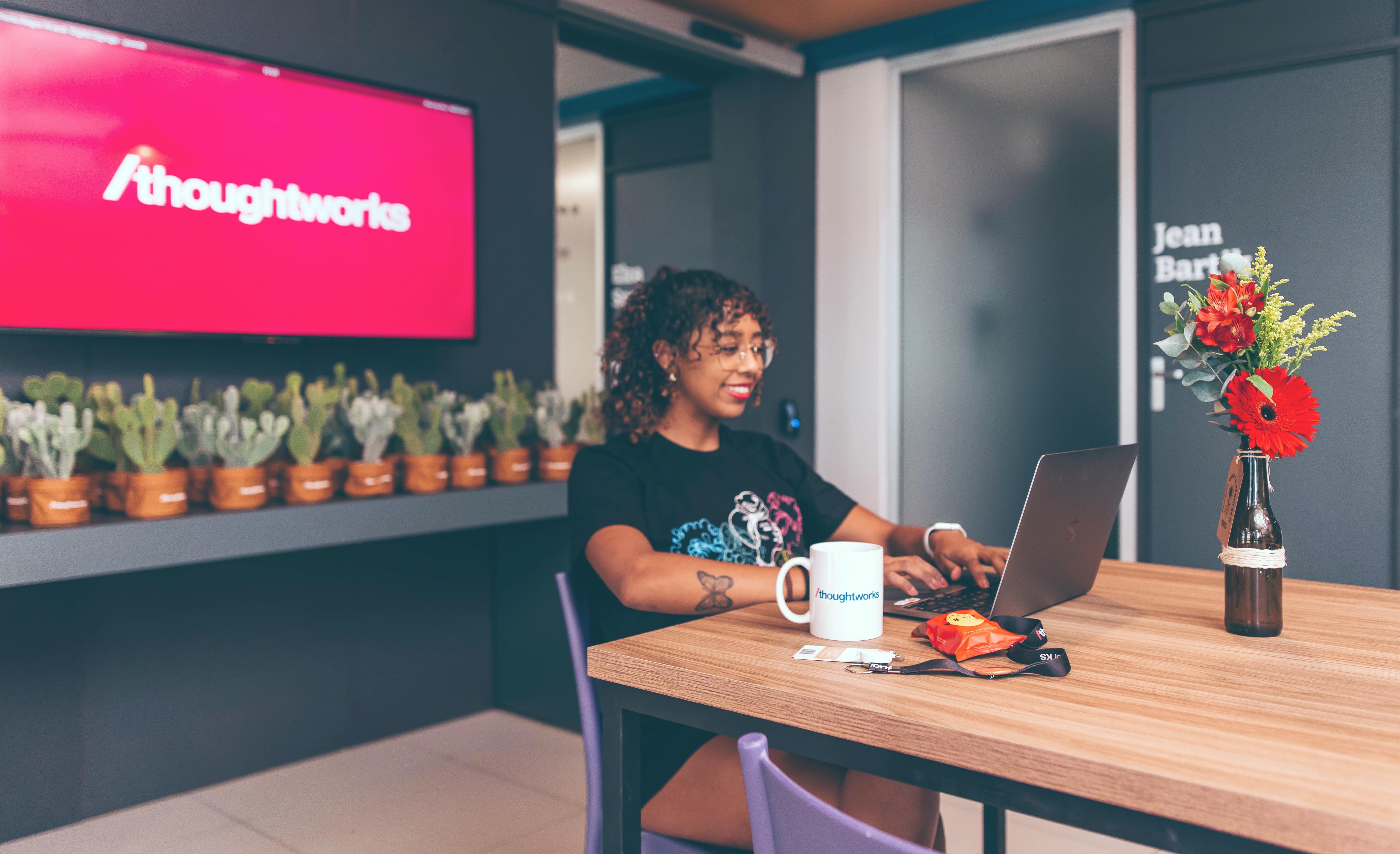 Una mujer sentada, frente a una mesa con su portátil y una taza de Thoughtworks. En el fondo, hay una televisión con el logo de Thoughtworks en la pantalla y plantas alrededor.