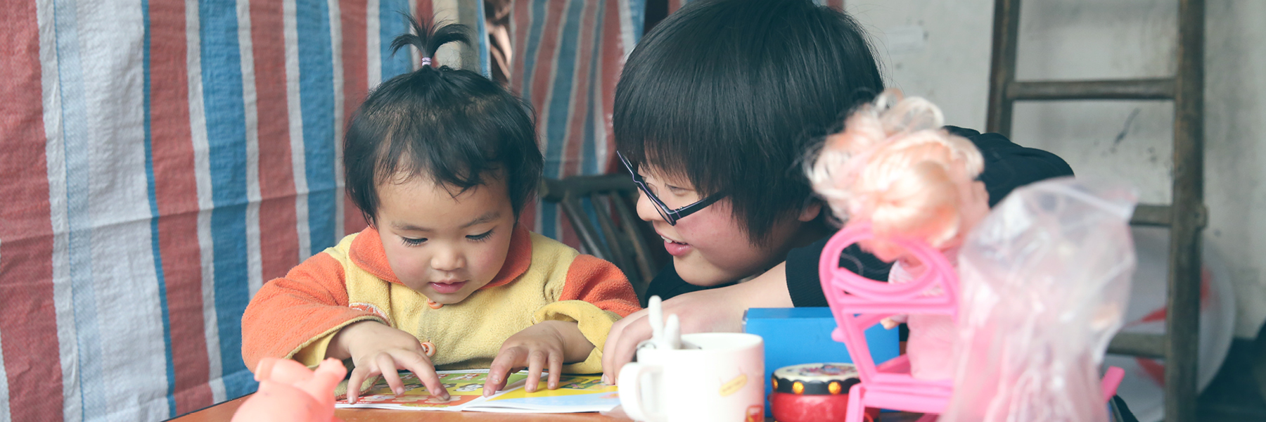 A young child and an adult both with black hair sit at a table, surrounded by childrens toys and reading from a piece of paper together 