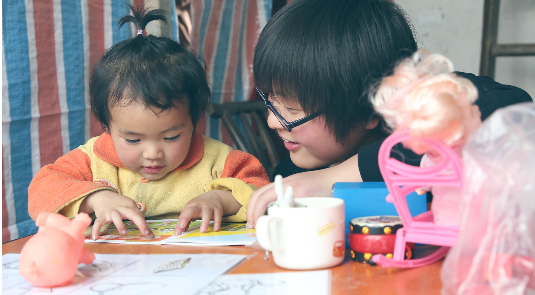 A young child and an adult both with black hair sit at a table, surrounded by childrens toys and reading from a piece of paper together 