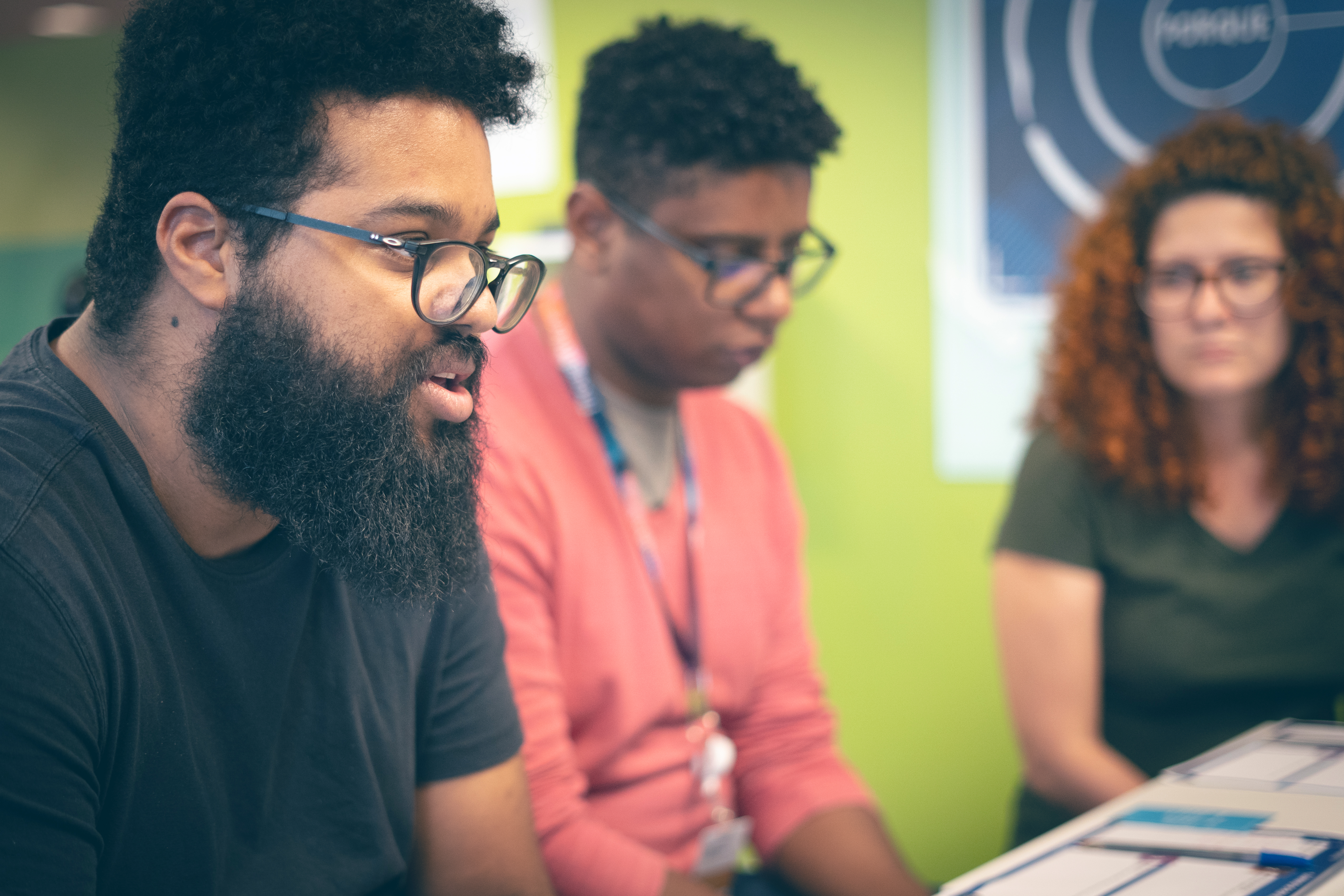 Two men and a woman can be seen in the frame. The man in front looks like he is talking to someone outside the frame. The group seems to be in a meeting room