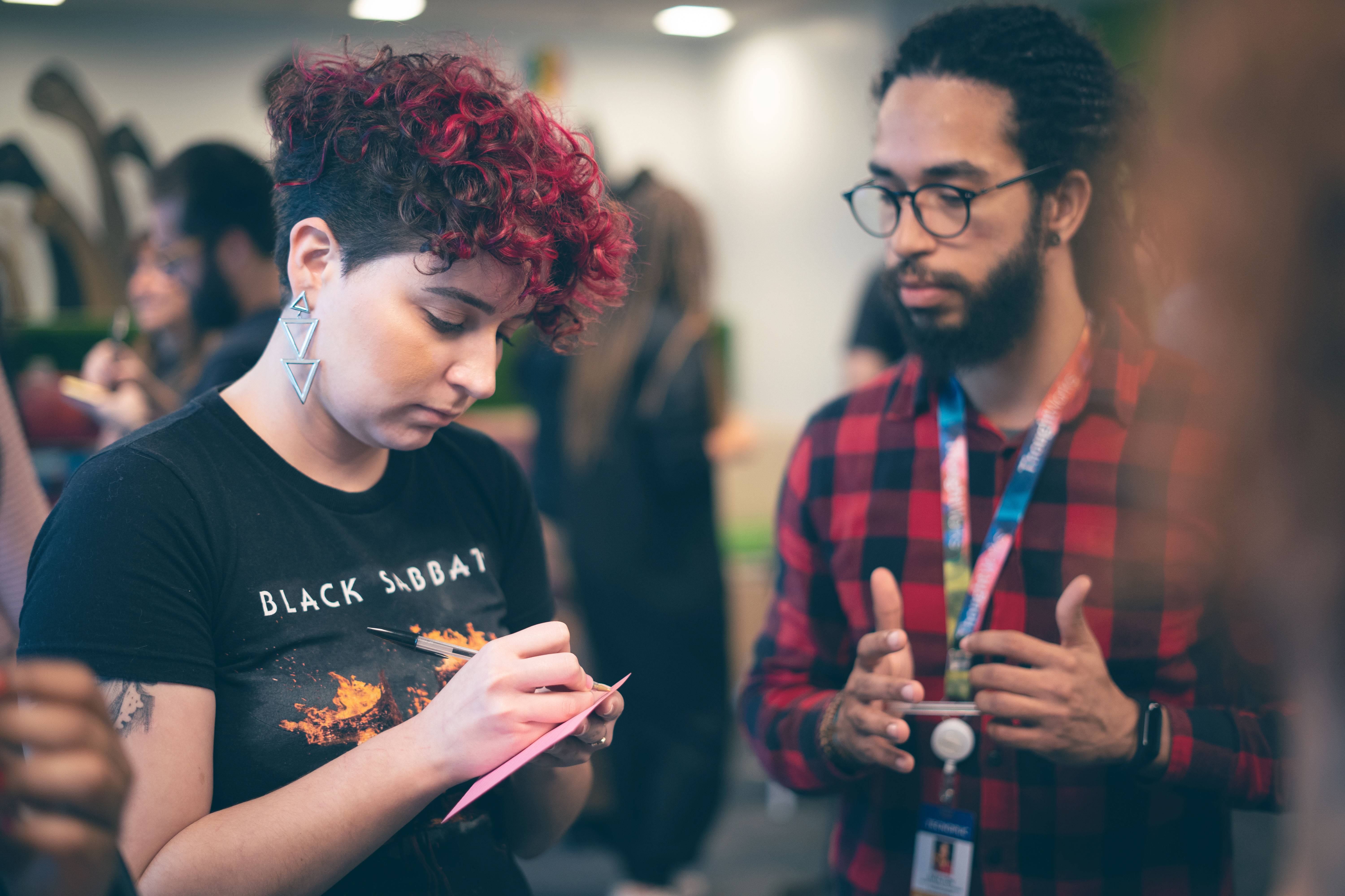 Woman with curly red highlights in a room of people. writing something on a card held in her hand. as a man with a beard looks on. 