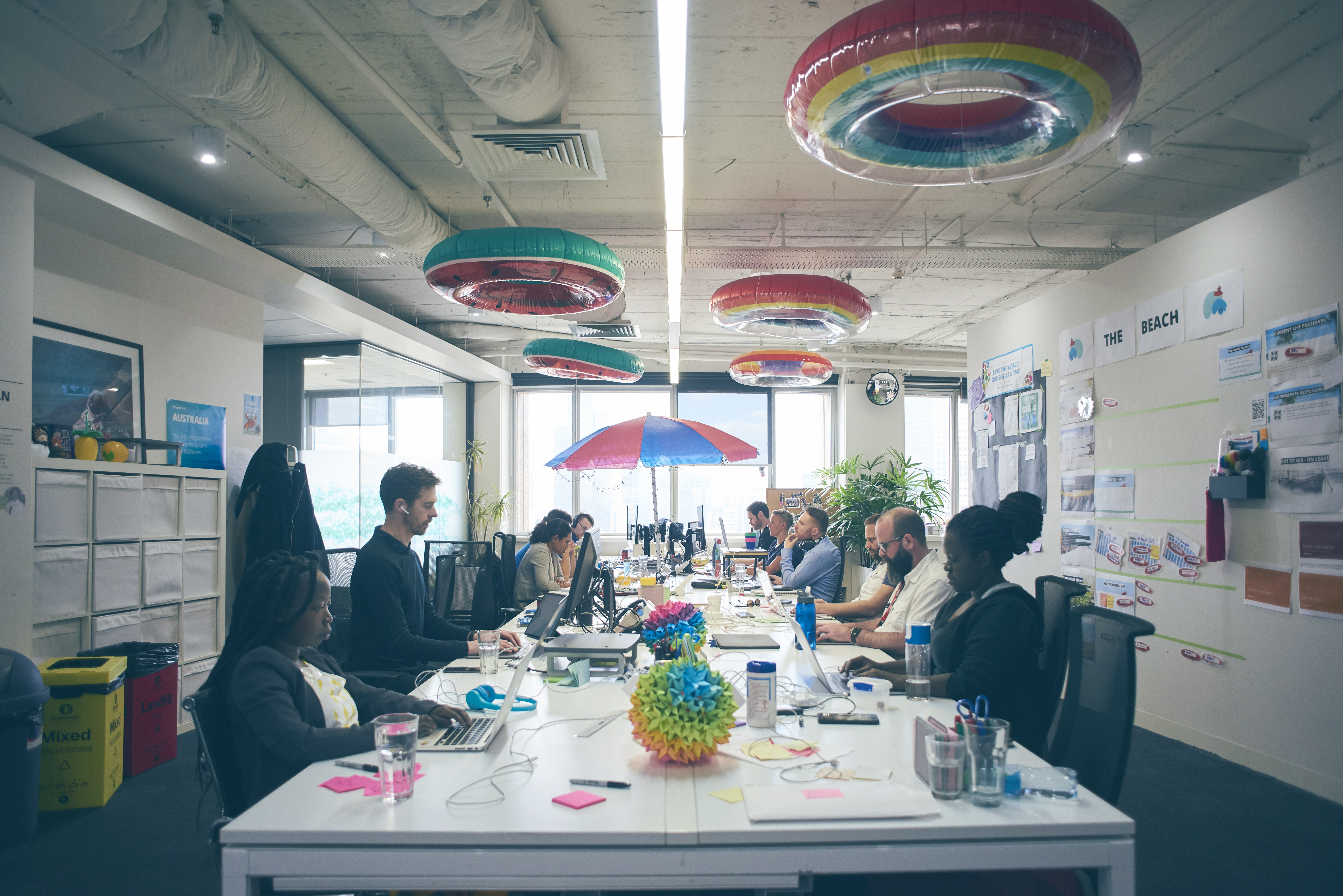 Thoughtworkers sentados alrededor de una mesa, trabajando. La oficina es vibrante, con hinchables de plástico multicolor en el techo.