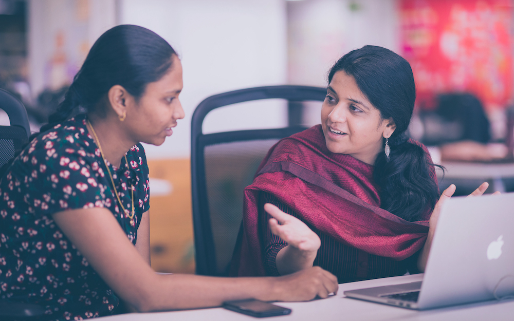 Photo of two women technologists sitting at a table at office and discussing a technical solution with each other