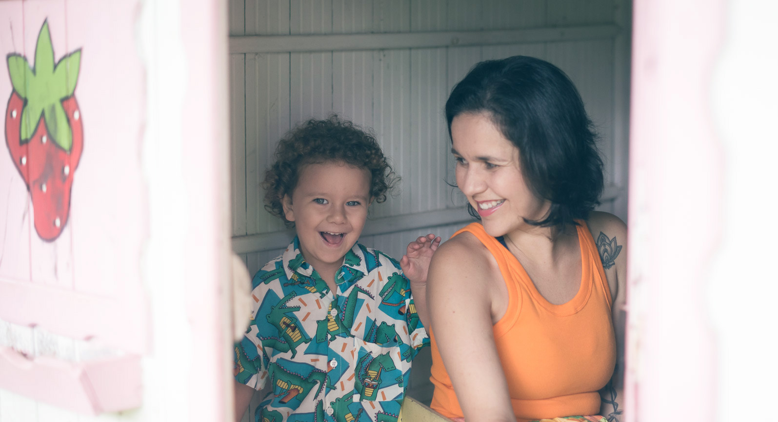 Marina Moscheta (Autora) sentada ao lado da sua criança dentro de uma casinha de madeira que possui um morango pintado do lado de fora.
