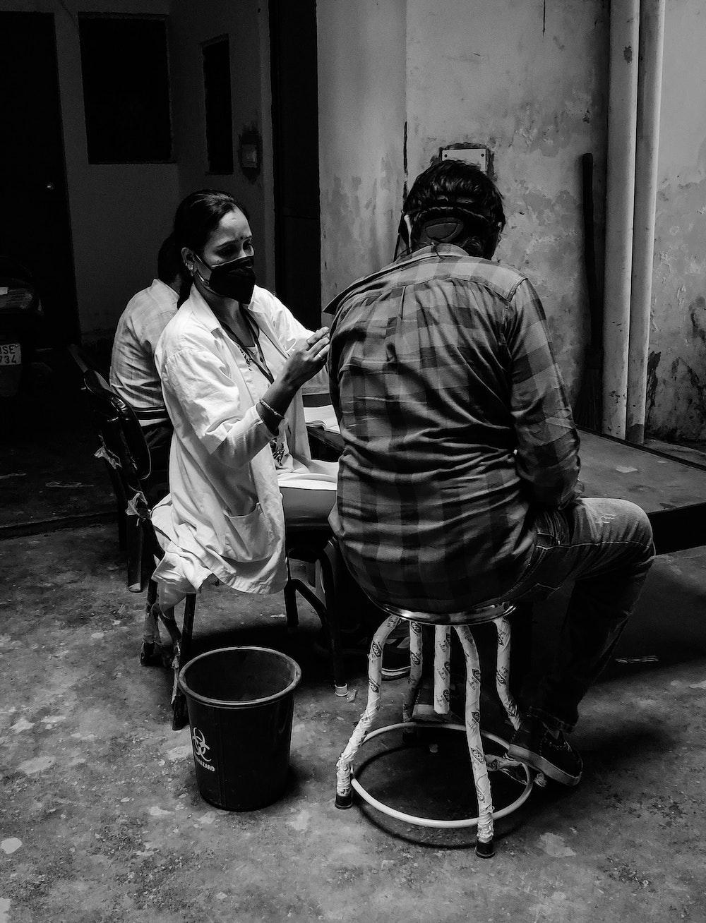 black and white picture of man sitting on a stool and a woman injecting him with a vaccine