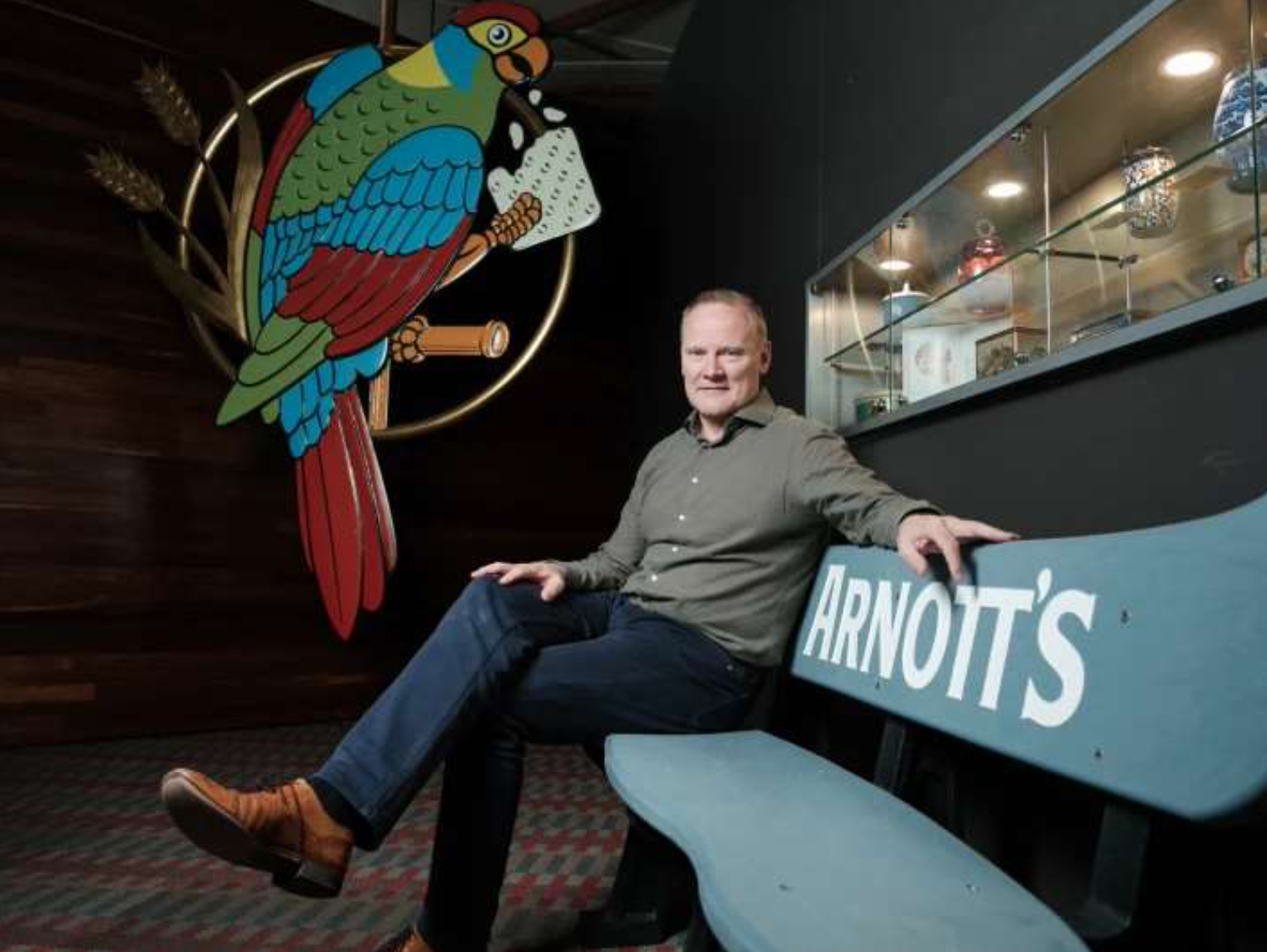 Simon Lowden sitting on a blue wooden bench with the word Arnotts painted in white. Next to Simon is a mobile with the macaw bird from the Arnotts logo.
