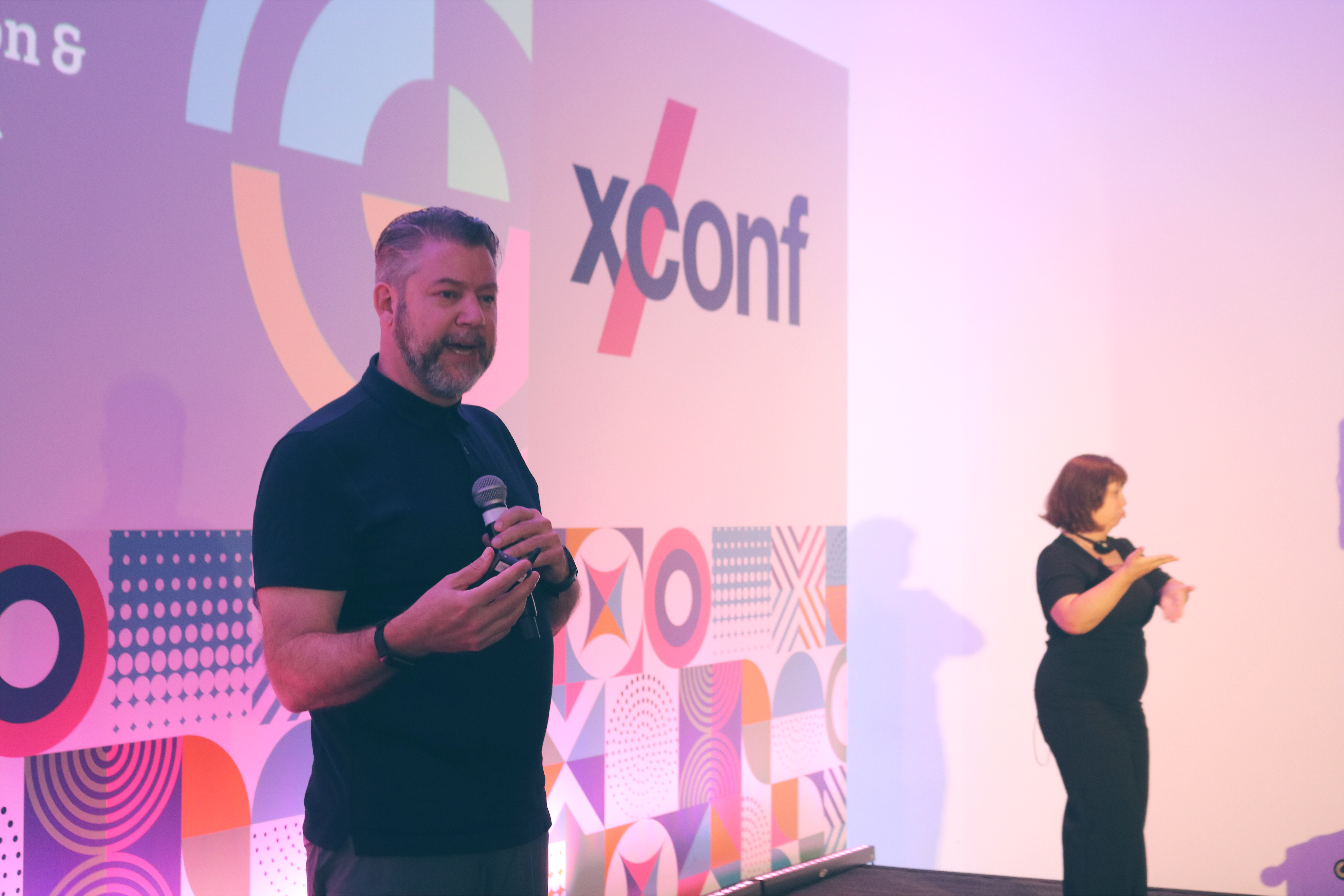 A speaker holds a microphone and presents on stage at XConf, with a colorful geometric backdrop featuring the XConf logo. Next to him, an interpreter provides sign language translation under soft pink and purple lighting. A speaker holds a microphone and presents on stage at XConf, with a colorful geometric backdrop featuring the XConf logo. Next to him, an interpreter provides sign language translation under soft pink and purple lighting.