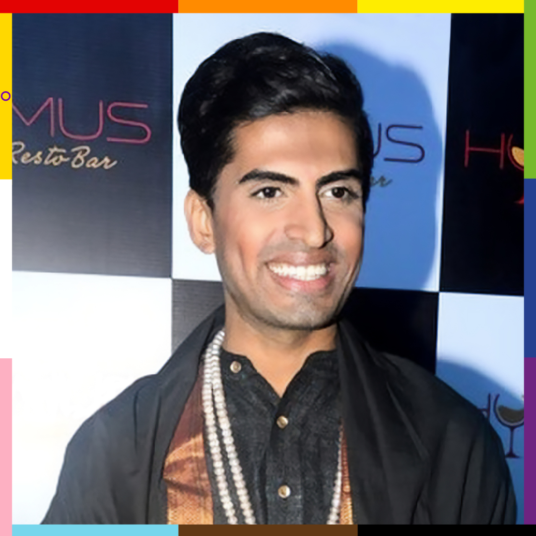 Sushanth, of Indian origin, is wearing a black kurta and a white beaded necklace stands smiling into a camera and framed with the Pride colors