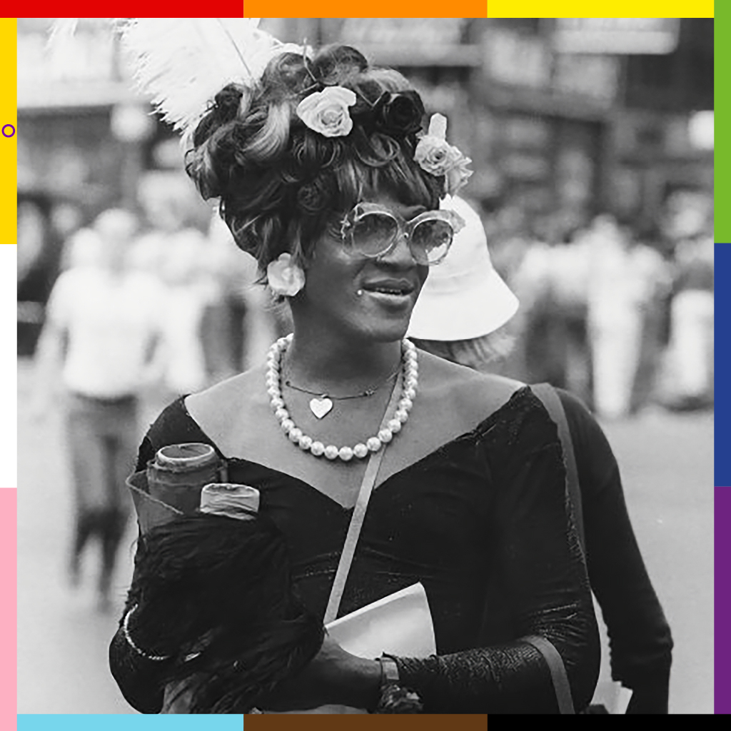 Marsha, a black woman in an elegant dress and sunglasses, with a hand-crafted progress pride frame