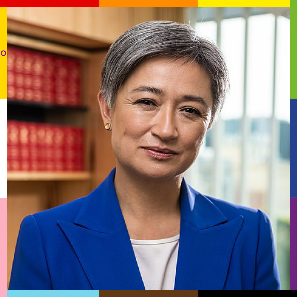 Penny, a woman with Malaysian heritage, in a blue suit with short hair and smiling eyes, is framed with the progress pride flag