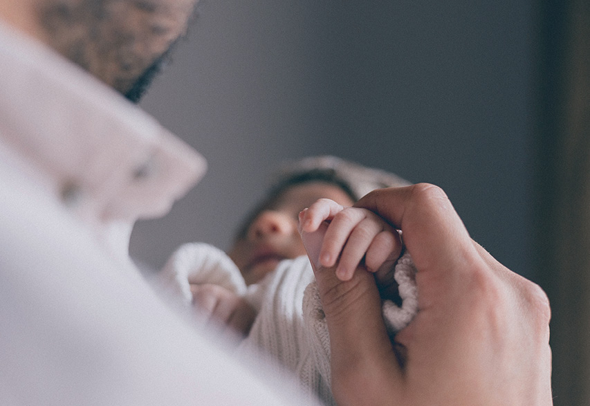 A tiny baby holding a hand. The man is in soft focus, and the baby wears a cotton hat and cardigan, looking very contented.