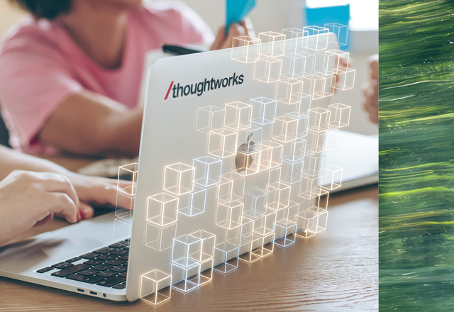 A close-up shot of a laptop featuring a Thoughtworks sticker on the lid, resting on a wooden table where Thoughtworkers are collaborating.