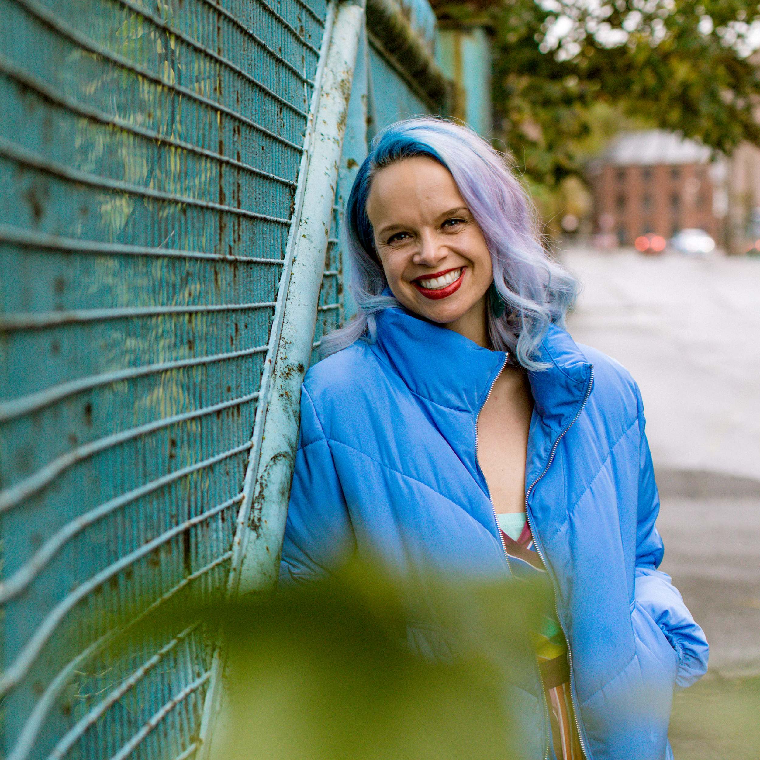 Amy, a Caucasian woman with vibrant blue and purple hair, wearing a blue jacket and smiling directly at the camera