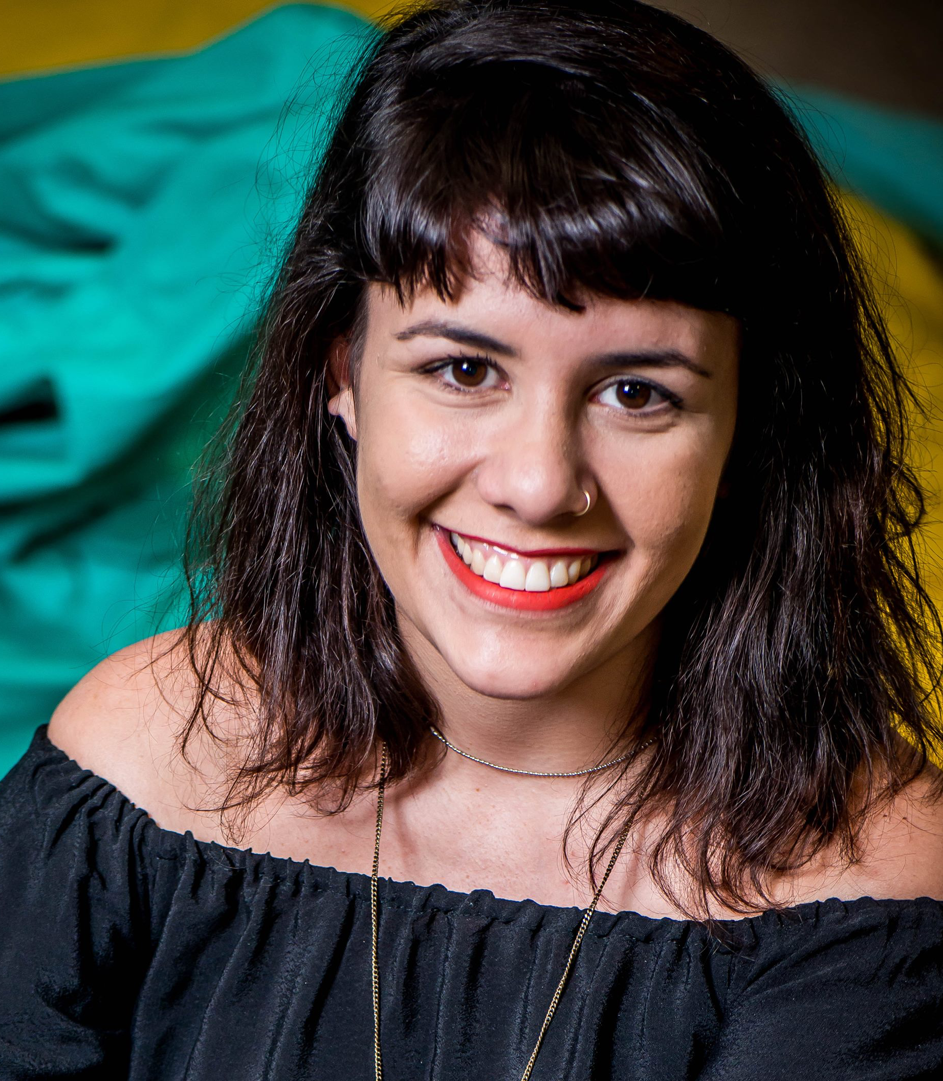 Headshot of Renata wearing a black off-shoulder top and red lipsitick,smiling into the camera. She has black hair in bangs and a nose ring.